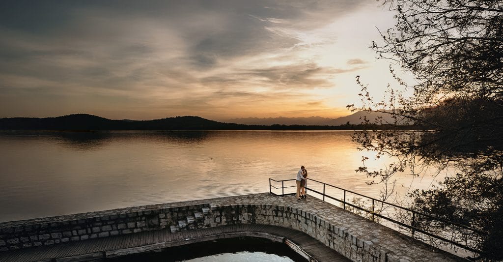 Engagement al Lago di Viverone di Ilaria e Luca. Sposi abbracciati sul molo di pietra con uno splendido tramonto sul lago di Viverone sullo sfondo. Foto aerea dal drone